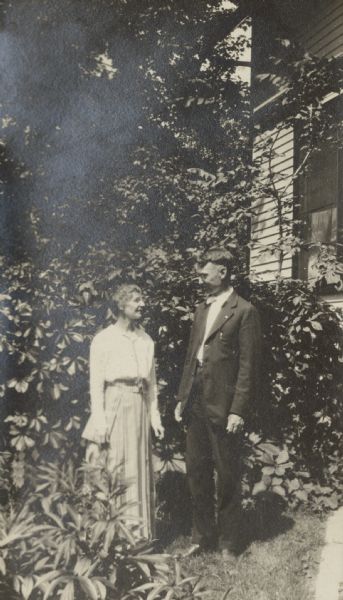 Lucy and W.A. Holt, wife and husband, are looking at each other while standing on the lawn with trees, shrubs, and a building in the background. Lucy Holt is holding a book in her right hand.
