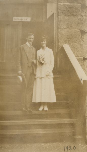 Wedding portrait of Madeleine Wood and Alfred Holt. They are standing close together on the stairs of a brownstone building, possibly a church or a court house. Above the open door to the building is a sign reading, "Entrance No. 1." Madeleine Wood is carrying a wedding bouquet. Alfred Holt is holding a straw boater hat. Caption reads: "New York, New York, July 15, 1920."


 