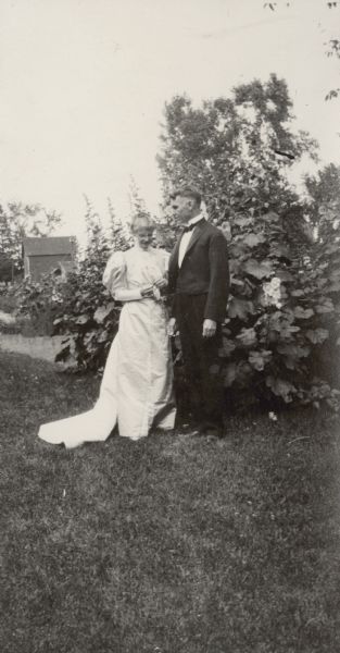 Marking their 25th wedding anniversary, Lucy and W.A. Holt are standing close together and posing for a portrait outdoors. Lucy Rumsey Holt may be wearing the wedding dress she wore in July 25, 1895. In the background are a shed, trees and shrubs. Caption reads: "25th Anniversary." 
