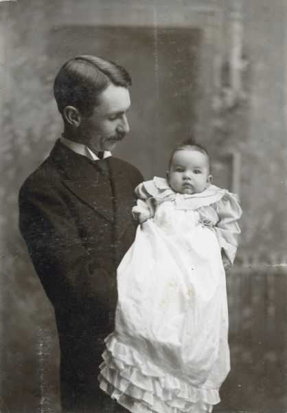 W.A. Holt is standing and holding his daughter Eleanor who is 3 months and 10 days old. She may be wearing a christening gown. This portrait was taken in a studio with a painted background. Caption reads: "Eleanor and Father, February 20th 1901, 3 months and 10 days." 