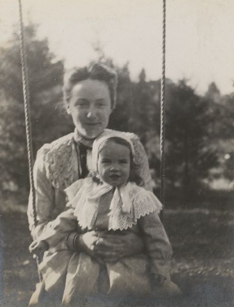 Minnie May Rumsey is sitting on a swing with niece Eleanor in her lap. There are trees in the background.