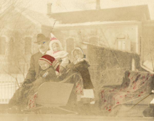 W.A. Holt and three children posing for a group portrait in the horse-drawn sleigh on Thanksgiving. Holding the reins, Alfred Holt is sitting next to his father in the front seat. W.A. Holt is holding younger daughter Eleanor Holt. Older daughter Jeannette Holt is standing up in the sleigh. Blankets cover the seats of the sleigh. A building is in the background. Caption reads: "A Thanksgiving Sleigh Ride. November 1903. Oconto."