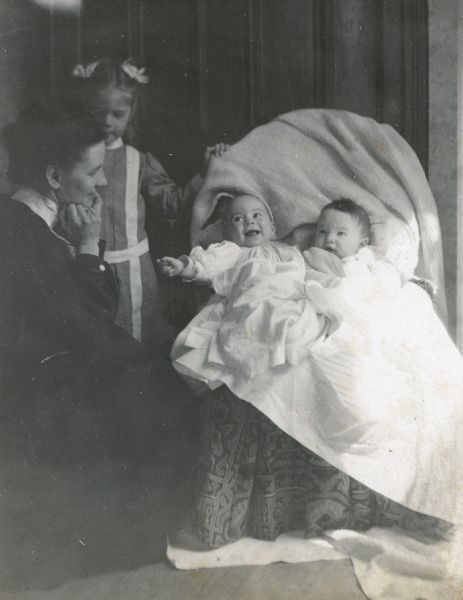 Little cousins Donald Holt and Juliet Stroh are sitting in the light, which is streaming through the window on the right. Lucy Holt or Juliet Stroh and the babies arelooking at each other. Either Harriet or Margaret Stroh is standing to the left in the background. Caption reads: "Juliet and Donald - A Visit to L.F."