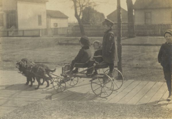 Fred Phelps is driving a small wagon with a team of dogs on the wooden sidewalk in Oconto. Eleanor and Alfred Holt are the passengers. Looking at the camera, Paul Garvey is standing on the sidewalk behind the wagon. Caption reads: "Now w're oxx!" Page caption reads: "Fred Phelps Team. Paul Garvey. E & A."
