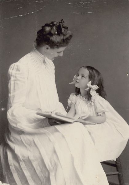 A studio portrait of mother (Lucy Rumsey Holt) and daughter (Eleanor Holt). Looking up, Eleanor Holt appears to be asking her mother to read her a story. They are both wearing dresses. Caption reads: "Read me a story mudder." Name listed is: Eleanor. Date listed on adjacent page is: 1903.

