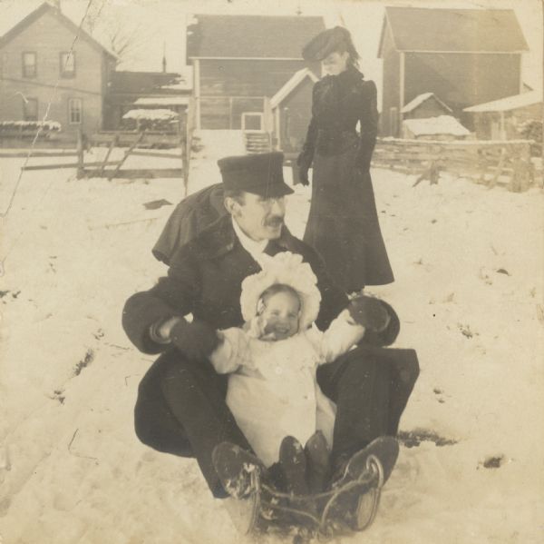 Smiling at the camera, daughter Eleanor Holt is tobogganing with her father W.A. Holt in the backyard of the Holt family home. Aunt Nell is standing behind the toboggan. There is a toboggan slide in the background. Note the steps on the right side of the toboggan slide for walking up to the top of the run. Caption reads: "Exercise After Thanksgiving Dinner 1903."