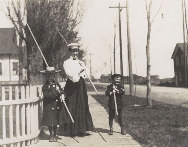 Miss McDonald, Jeannette Holt, and Alfred Holt are going fishing. Standing on the wooden sidewalk, they are holding long bamboo fishing poles over their shoulders. Caption reads: "Miss McDonald." Date reads: "Spring 1904." Nearby caption reads: "Jeannette and Alfred going fishing with 'Teacher.'"