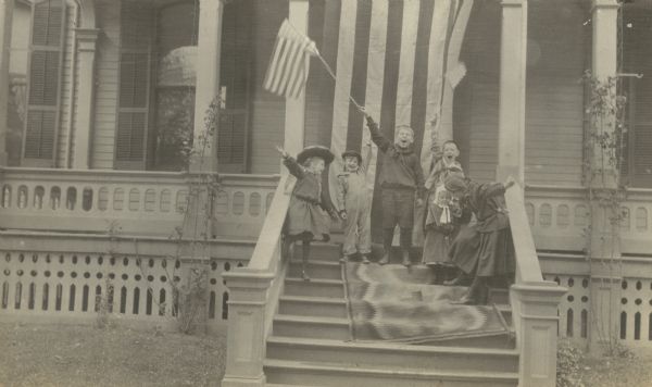 A group of children are standing on a carpet over the front steps of the house. Two boys are waving flags. A large flag is hanging from the second story. The girl on the far left may be Jeannette Holt, and the boy next to Jeannette Holt may be Alfred Holt. The little girl on the right may be Eleanor Holt. The home's wraparound porch is in the background. Caption reads: "Hurrah for the 'Red white and blue.'" Page caption reads: "Lake Forest 1904." 