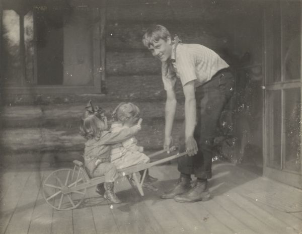 Joe Rumsey (?) is pulling the wooden cart holding cousins Donald Holt and Juliet Stroh. They are playing on the porch at Island Lodge. Caption reads: "Joe as 'baby tender.'"
