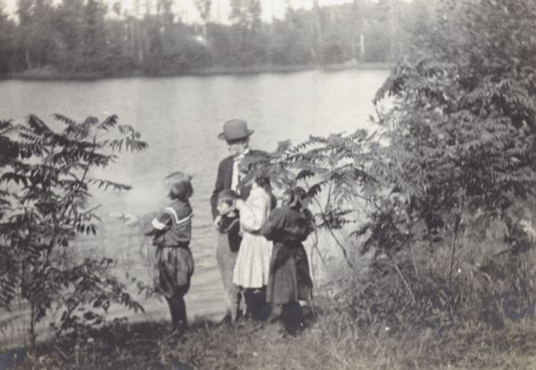 Harriet Stroh is taking a shooting lesson from her grandfather Captain I.P. Rumsey, a Civil War veteran. Alfred Holt is standing in front of his grandfather. Jeannette Holt is standing to the right of Alfred Holt. And Margaret Stroh is standing to the right of Jeannette Holt. The group is on the shore of Archibald Lake. Caption reads: "Lesson in Shooting - Grandfather instructor." Page caption reads: "Lakewood. August 1905."