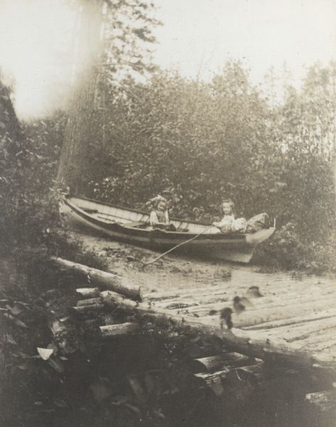 Using a long wooden stick as an oar, Donald Holt is rowing the boat, which is beached on land. Juliet Stroh is sitting in the boat with Donald Holt. A log bridge is in the foreground. Caption reads: "First lesson in rowing."