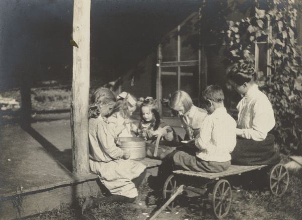 Children are shelling lima beans on the porch at Island Lodge. Names from left to right on the porch: Jeannette Holt, Eleanor Holt, Donald Holt, and Harriet or Margaret Stroh. Sitting on the wooden wagon, from left to right: Alfred Holt and {Unknown}. Caption reads: "Lima Beans for Dinner." 