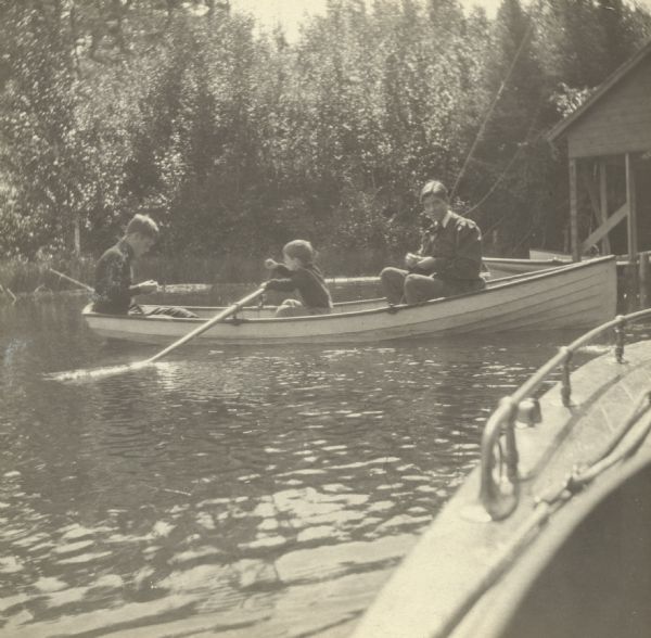 Alfred Holt (center) is rowing the boat on Archibald Lake while Richard Rumsey (?) and James McClure are baiting hooks for fishing. The wild shoreline and the Holt boathouse are in the background. The Islander motorboat is in the foreground. Caption reads: "Fishing Party." Names, from left to right, are: Richard, Alfred, James."