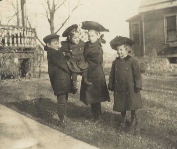 Donald Holt, age 4, is being carried by Alfred Holt and Jeannette Holt. This was the day that Donald Holt had his curls cut. Eleanor Holt is standing next to Jeannette Holt. They are in the front yard of their family home in Oconto. Caption reads: "The Quartette, Oconto, November 1905."