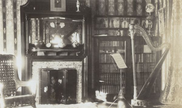 Lucy Rumsey Holt's concert harp and music stand are on the right side of the living room or music room. Behind the harp is a large wooden bookcase with glass doors, filled with leather bound books. Above the bookcase is a framed work of art, possibly a pre-Raphaelite painting. In the center is a fireplace and mantel, with a lacquer screen in front of the fireplace. Above the fireplace is a mirror, reflecting a chandelier. To the left of the fireplace is a wicker or rattan chair.