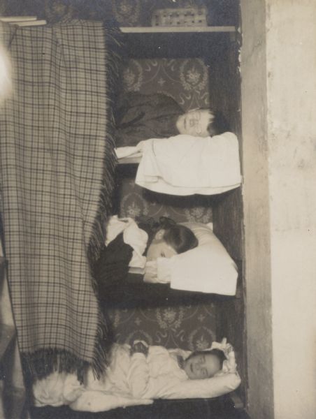 Three Holt children are pretending to be sleeping in the sleeping car of a train, with closet shelves as the train beds. A wool blanket with fringe is the privacy curtain. On the left are a set of stairs or a ladder. Caption reads: "Playing sleeping car in the closet on the stairs." Names from upper bunk to lower bunk: "Alfred Holt, Jeannette Holt, and Eleanor Holt."