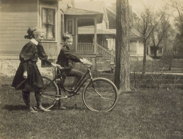 Alfred Holt is jumping on his new bicycle in the yard. Jeannette Holt is resting her hand on her brother's bicycle seat. Neighboring homes are in the background. Caption reads: "Alfred's New Wheel." 