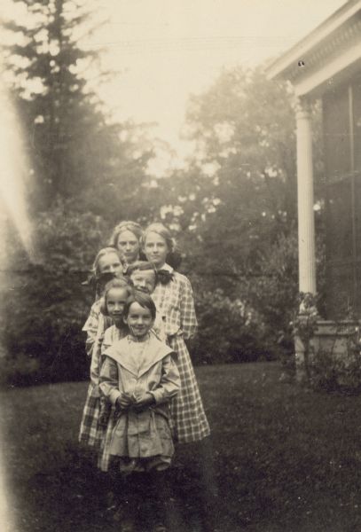 The Holt and Stroh children are posing for a group portrait in the yard of "The Evergreens," home of Lucy Rumsey Holt's parents. Names from back to front: Margaret Stroh, Harriet Stroh, Jeannette Holt, Alfred Holt, Eleanor Holt, and Donald Holt. Caption reads: "The Children's Ladder. M,H,J,A,E,D." 