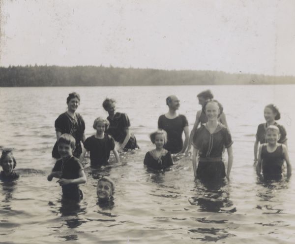 A group of women, men, and children are bathing in Archibald Lake. Caption reads: "The more the merrier. Mrs. Sidley, Juli, Mr. Sidley, and Arch." 