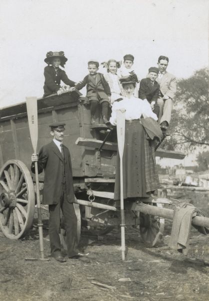 A group of Holts and Macartneys is posing for a photograph after completing a trip down the river. Standing next to the wagon, W.A. Holt is holding an oar. Mrs. Macartney, leaning on an oar, is preparing to step down. Sitting in the wagon, names from left to right: Jeannette Holt, Donald Holt, Eleanor Holt, Arthur Macartney (?), Alfred Holt, and Rev. John Robertson Macartney." Caption reads: "After a trip down river. Mr. Macartney, Mrs. Macartney, Arthur, and the Holts."


