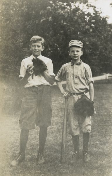 Nathan McClure (l) and Alfred Holt (r) are dressed to play baseball. McClure is wearing a baseball glove and holding the ball. Holt is leaning on the bat. His baseball glove is hanging from the belt loop on his knickers. Caption reads: "Baseball 'Fans' Nathan and Alfred."