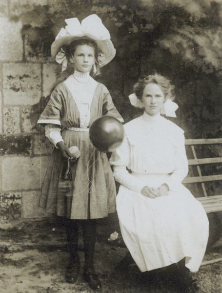 Jeannette Holt is standing and holding something that looks like a ballon on a string. Next to Holt, cousin Margaret Stroh is sitting with her hands in her lap on the bench. A stone wall is in the background. Caption reads: "Jeannette, Margaret. The Guild Bazaar." 