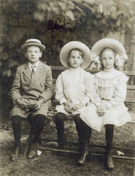 Three Holt children are sitting on a park bench in front of a stone wall. From left to right: Alfred Holt, Donald Holt, and Eleanor Holt. Alfred Holt appears to be holding a small wooden sailboat toy. Caption reads: "The Guild Bazaar. Alfred, Donald, Eleanor." 