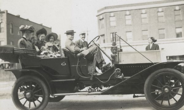 Looking at the camera, Joe Rumsey is sitting in the driver's seat of his sedan. The auto has lights and a horn. Morris Rumsey is at his brother's side. In the back seat, left to right, Julie Stroh (?), Elizabeth Stroh with doll, Aunt Dena, and Juliet Unknown are sitting next to each other. Standing on the street, between the auto and the building, is Harriet Stroh.

Caption reads: "Joe's Auto. Julie and Juliet leaving for Woodhull." In back seat, from left to right, "Julie - Elizabeth, Aunt Dena - Juliet." In front seat, from left to right: "Joe, Morris." Standing in front of the auto: "Harriet." 