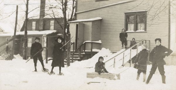 Children are playing winter sports, including sledding and snowshoeing, in the backyard. The slide and sleds are made of wood. The snowshoers are holding hockey sticks. On the right, Alfred Holt is holding the sled rope, between the sled and the boy standing akimbo.
