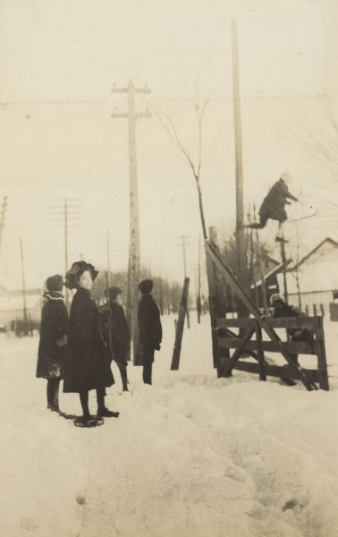 On a snowy day, Alfred Holt is leaping from the gate post. Several children are watching him. A girl, wearing a floppy winter hat, is looking at the camera. There are utility poles in the background.