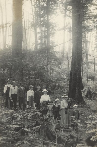 Slightly elevated view of a group of men, women, and children on the blackberry trail. Alfred Holt is first in the line, followed by his mother Lucy Rumsey Holt. Bringing up the rear is Arthur D. Wheeler, using a walking stick. Many in the group are carrying a pail or cup for collecting blackberries. Caption reads: "Blackberry Trail. By Mr. Tiers."