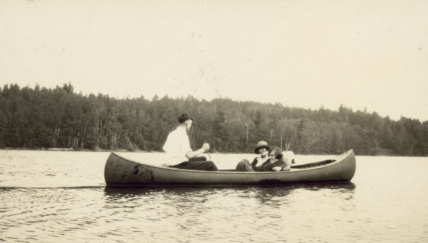 Wearing a hat, Madeleine Wood Holt is smiling, sitting next to Helen Wood, on the floor of the canoe in Archibald Lake. With his back to the camera, Alfred Holt is paddling. Forest and shoreline are in the background. Caption reads: "Alfred, Madeleine, and Helen Wood." 