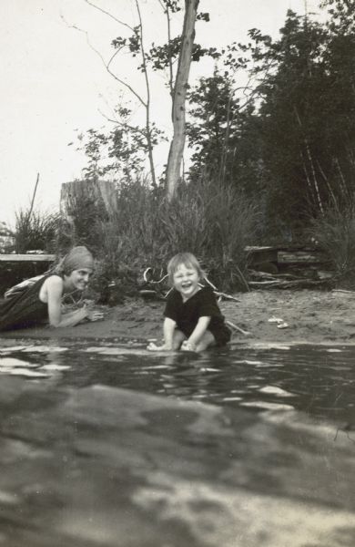 View from Archibald Lake of toddler Barbara Holt who is sitting on the beach and laughing. Her feet are in water. Aunt Jeannette Holt, wearing a headscarf, is watching her niece Barbara. Caption reads: "J.R.H. and Barbara."