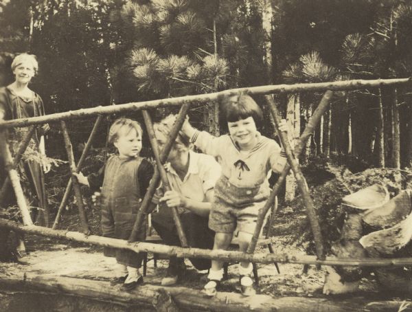 Arthur Holt (far right) is pushing his head through the hemlock gate. Sister Barbara is watching him. Lillian Wheeler (?) is looking at Barbara. A small wooden chair is behind each child. Lucy Rumsey Holt is walking in from the left. Split logs are on the far right. Pine trees are in the background.