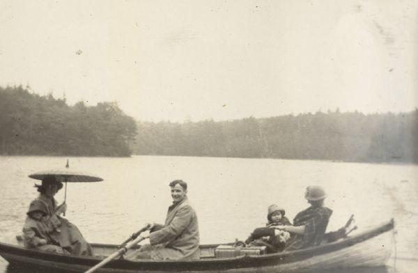 With hands on the oars, Alfred Holt is looking at the camera as he and his family depart the Island after a vacation on Archibald Lake. On the left is Madeleine Wood Holt, under an oil-paper umbrella, with son Arthur. On the right is an unidentified woman and Barbara Holt. There is one suitcase in the boat. The forest surrounding Archibald Lake is in the background. Caption reads: "A rainy departure."