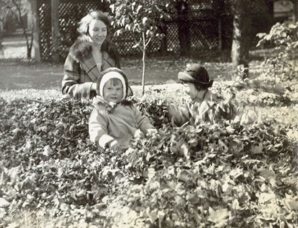 Barbara Holt, on the left, is looking at the camera while sitting in a pile of dried leaves. Behind her is Aunt Eleanor Holt, who is wearing a fur-collared wool coat. Aunt Eleanor is looking at Arthur Holt who is on the right playing in the leaves. Caption reads: "Eleanor, Barbara, and Arthur."