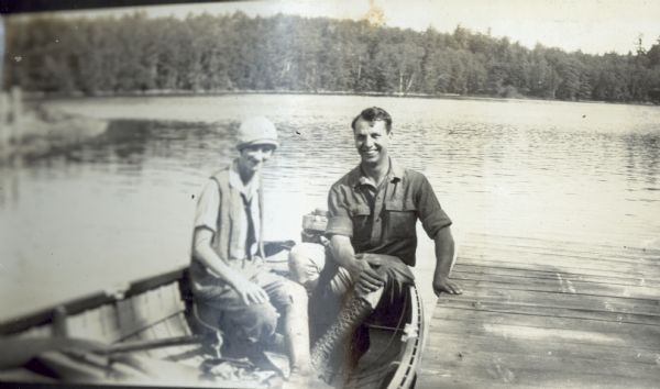 Judy Unknown and Pete Blanchard are sitting in the boat at a dock on Archibald Lake. Pete is wearing lace-up leather work boots. There appears to be an outboard motor on the back of the boat. In the background is Archibald Lake shoreline and forest. Caption reads: "Judy and Pete."