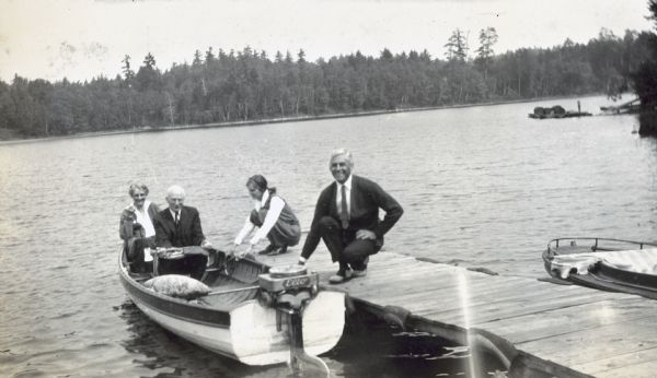 View toward a man, sitting on the middle bench in the motor boat, holding an axe on his knees. A woman, possibly Anna Holt Wheeler, is sitting on the back bench. Jeannette Holt is holding the boat at the pier. Mr. Garrison, smiling at the camera and kneeling on the pier, is also holding the side of the boat. The company name "Elco" is painted on the boat's motor. 

The forested shoreline of Archibald Lake is in the background. The Holt Lumber Company never harvested the old-growth trees along this shoreline.

The back side of the photograph reads: "Wheeler, Garrison."