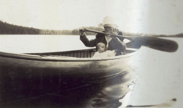 Lucy Rumsey Holt is paddling the canoe on Archibald Lake with grandson Peter DeWitt in her lap. There is a forested shoreline in the background. Caption reads: "Grandma Holt and Peter DeWitt."
