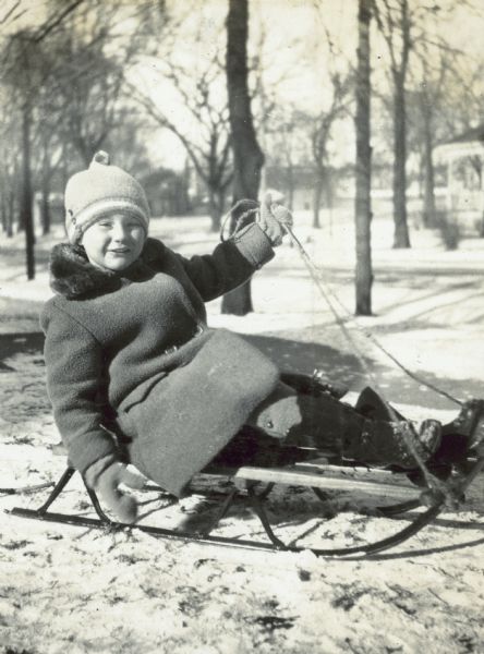 On a sunny winter day, Barbara Holt is ready to go sledding. She is sitting on the wooden sled and holding the rope in her left hand.
