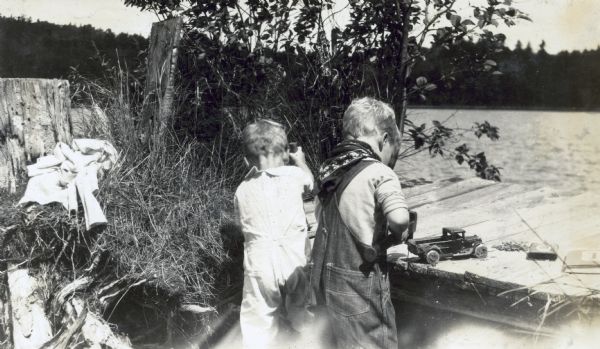 With their backs facing the camera, Peter DeWitt (l) and Douglas DeWitt (r) are standing side by side, hammering nails into a pier on Archibald Lake. Douglas DeWitt is wearing denim overalls with a bandana around his neck. There are several items on the pier: a sandy toy truck, some nails, a small can, and a book.
