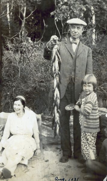 Standing on a sandy beach on the Island at Archibald Lake, Grant Stroh is holding up a string of fish. To the left of Stroh, a young woman, possibly his daughter, is sitting on a beach towel, leaning up against a big log. To the right of Stroh, a girl, possibly one of his grandchildren, is holding a garden trowel.

