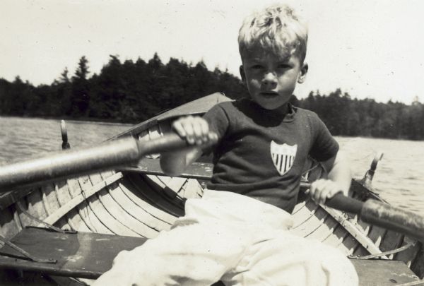 Douglas DeWitt, one of the Holt grandchildren, is rowing on Archibald Lake with an island in the background. 