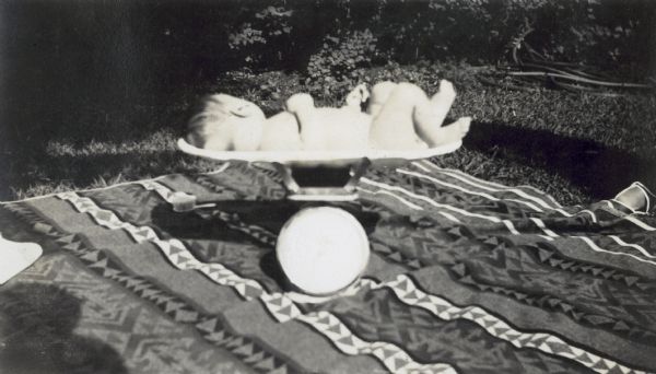 A baby, probably Donald Holt, Jr., is being weighed on a small scale outside. There is a blanket, with a geometric pattern, underneath the scale.