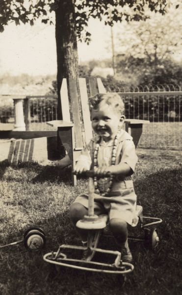 Wearing a necklace of wooden beads. Donald Holt, Jr. is riding a 4-wheel cycle in the yard. Behind him is an Adirondack Chair, a tree, a sandbox, and a wire fence.