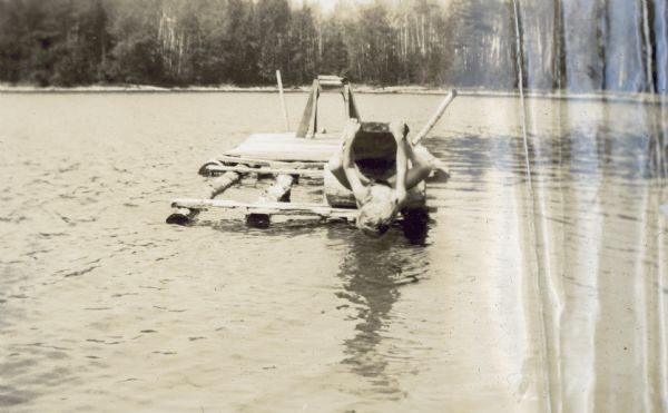 A boy is hanging upside down from the diving raft on Archibald Lake. Similar to many items owned by the Holt family, this raft probably was made by hand with local wood.