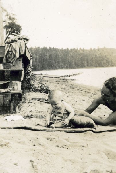A baby boy, probably Robert James DeWitt, is playing on a sandy beach. A woman, probably his mother Eleanor Holt DeWitt, is with him.