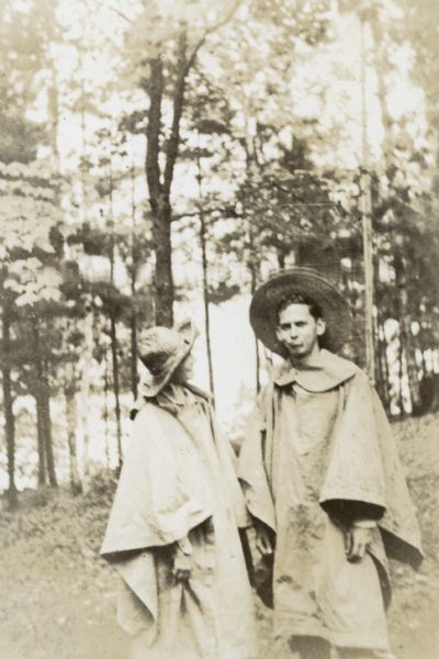 A man and a woman, probably Eleanor Holt DeWitt and Donald Soule DeWitt, are standing together, wearing rain ponchos and wide-brimmed hats. Trees and Archibald Lake are in the background.