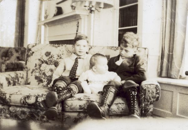 From left to right: Peter DeWitt, baby Robert Dewitt, and Douglas DeWitt sitting on a couch. The two older boys are wearing lace-up leather work boots.