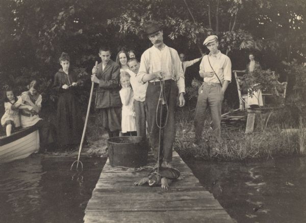 View towards Mr. Ferguson who is standing on a pier holding a rope with either a mud turtle, or a snapping turtle, tied to it. Behind him on the shoreline is a group of people. Among them, second from the left, is Jeannette Holt sitting on the side of a boat. A young man, on the left, is leaning on a pitchfork, with the tines in the lake. Wearing glasses, Donald Holt is standing directly behind Mr. Ferguson. Lillian Wheeler may be standing to the left of Donald Holt. Wearing a cap, Henry Thurston is standing on shore, on the right side. Lucy Rumsey Holt is standing at the far right, near the bench. Caption reads: "Mr. Ferguson and the Turtle." 